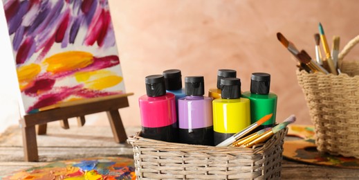 Photo of Acrylic paints of different colors and brushes in wicker basket on wooden table