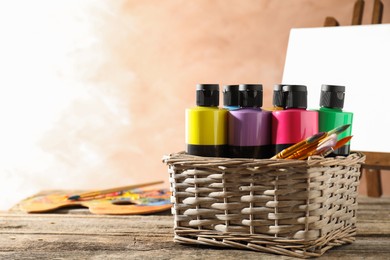 Photo of Acrylic paints of different colors and brushes in wicker basket on wooden table, space for text