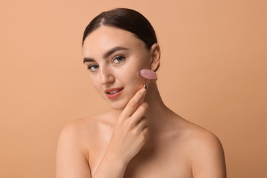 Beautiful young woman doing facial massage with roller on beige background