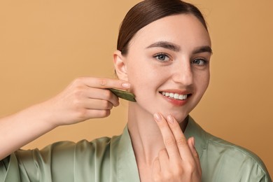 Beautiful young woman doing facial massage with gua sha tool on beige background