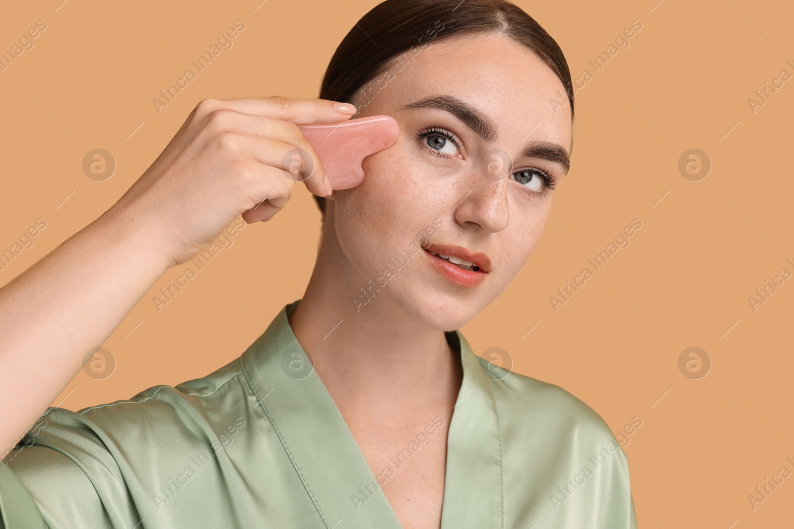 Photo of Beautiful young woman doing facial massage with gua sha tool on beige background