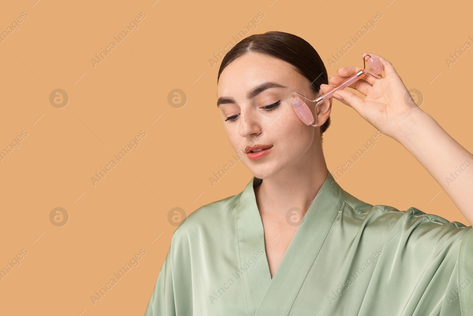 Photo of Beautiful young woman doing facial massage with roller on beige background, space for text