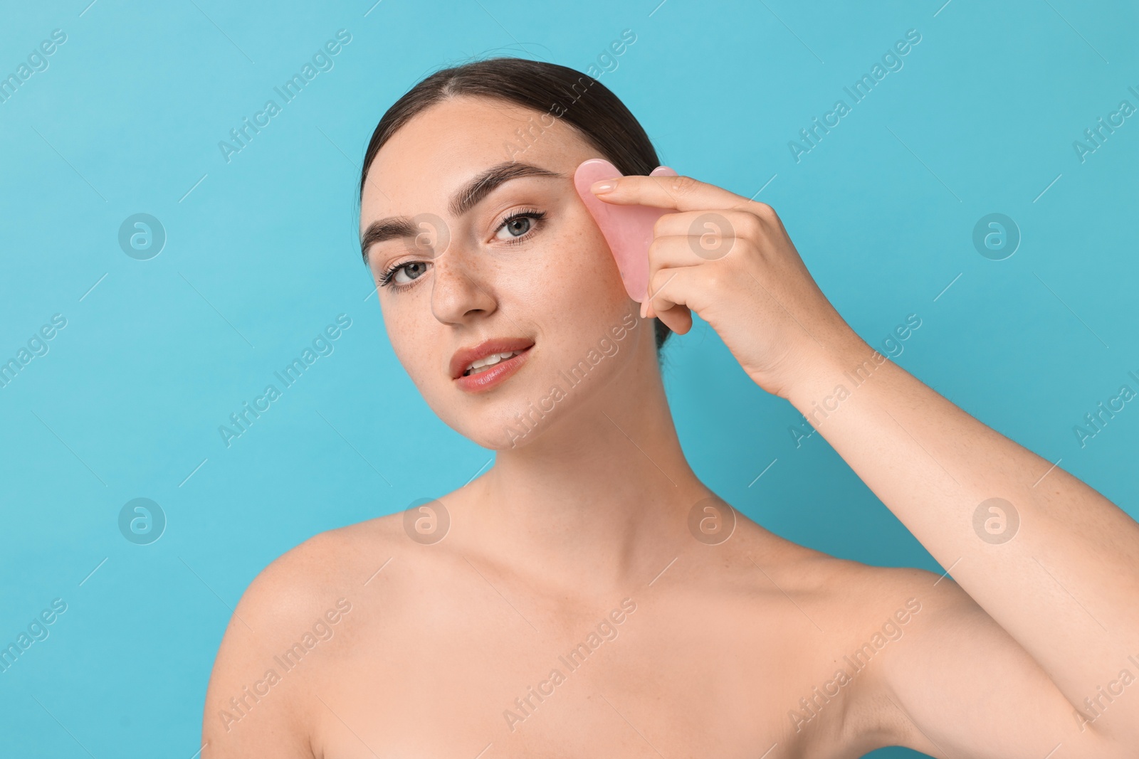 Photo of Beautiful young woman doing facial massage with gua sha tool on light blue background