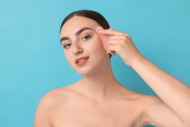 Beautiful young woman doing facial massage with gua sha tool on light blue background