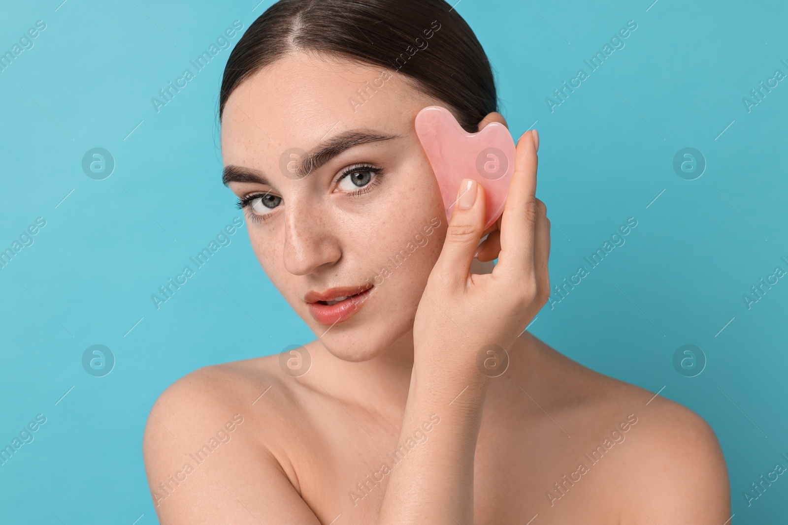 Photo of Beautiful young woman doing facial massage with gua sha tool on light blue background