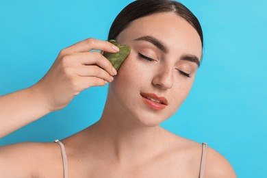 Photo of Beautiful young woman doing facial massage with gua sha tool on light blue background