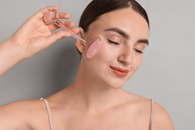 Beautiful young woman doing facial massage with roller on grey background