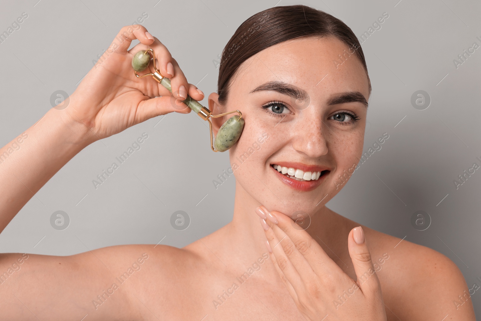 Photo of Beautiful young woman doing facial massage with roller on grey background