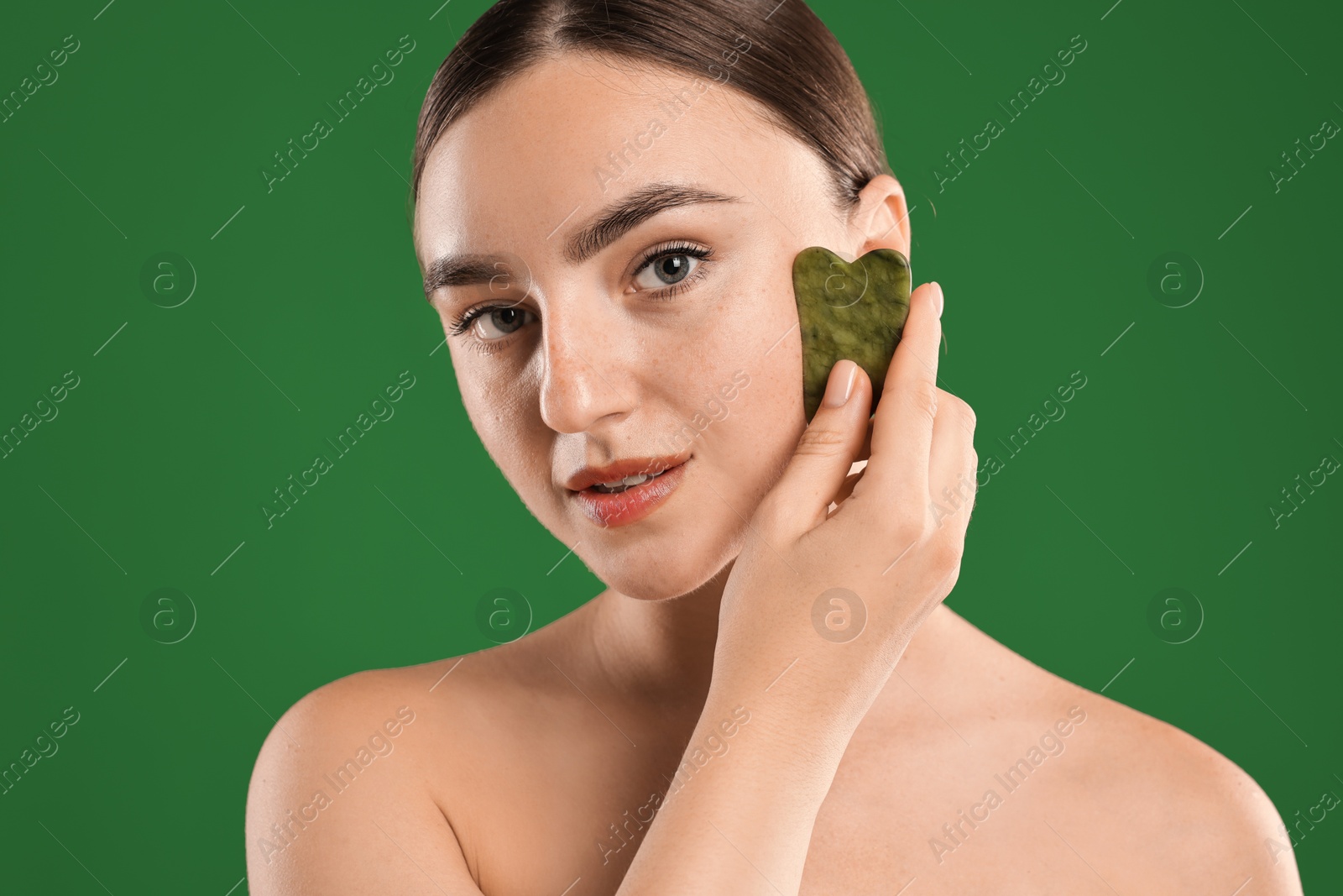 Photo of Beautiful young woman doing facial massage with gua sha tool on green background