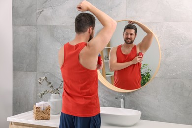 Photo of Smiling man applying solid deodorant near mirror at home, back view