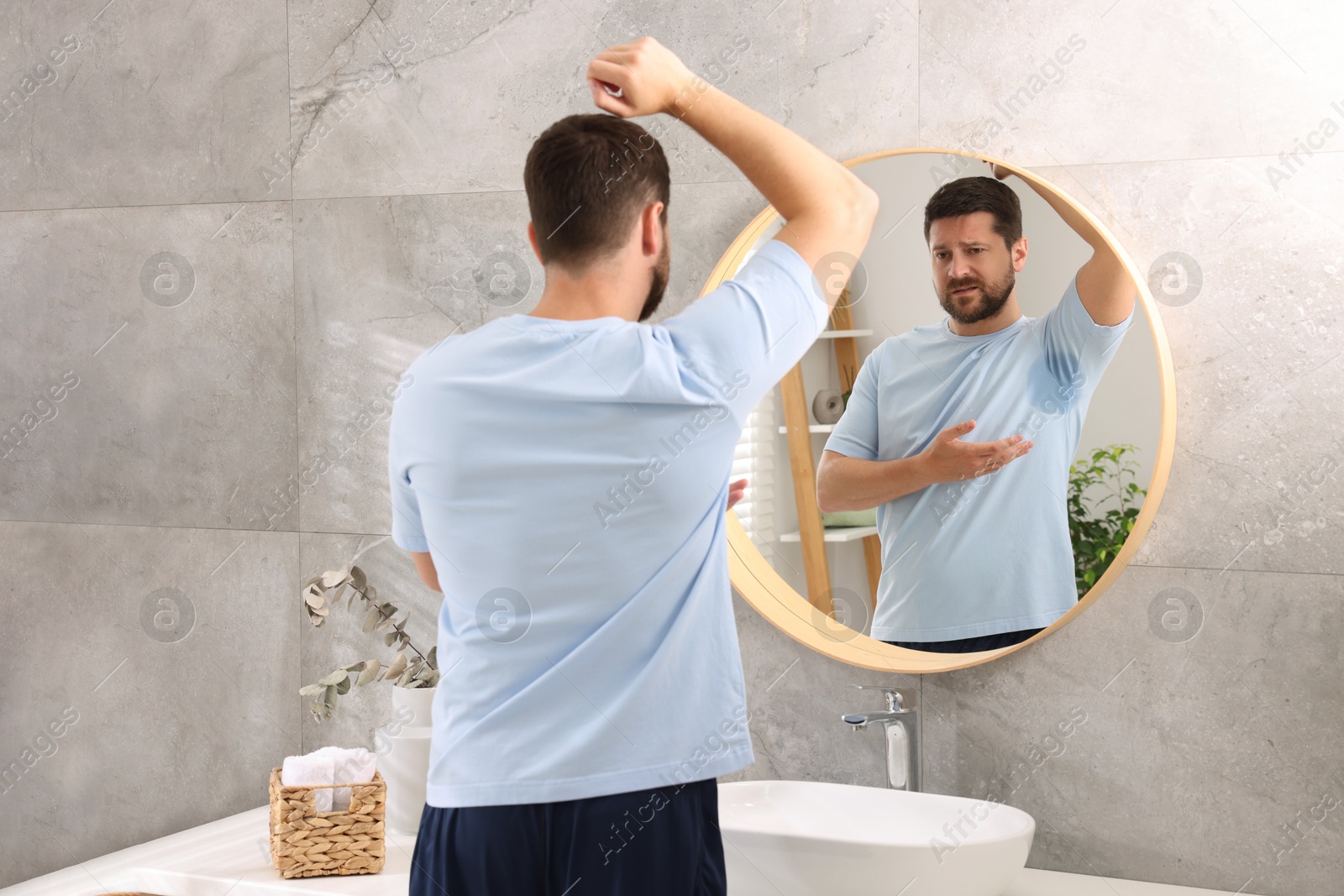 Photo of Emotional man in t-shirt before using deodorant near mirror at home, back view