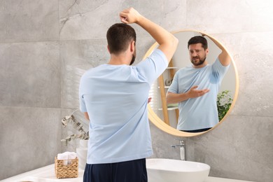 Photo of Emotional man in t-shirt before using deodorant near mirror at home, back view