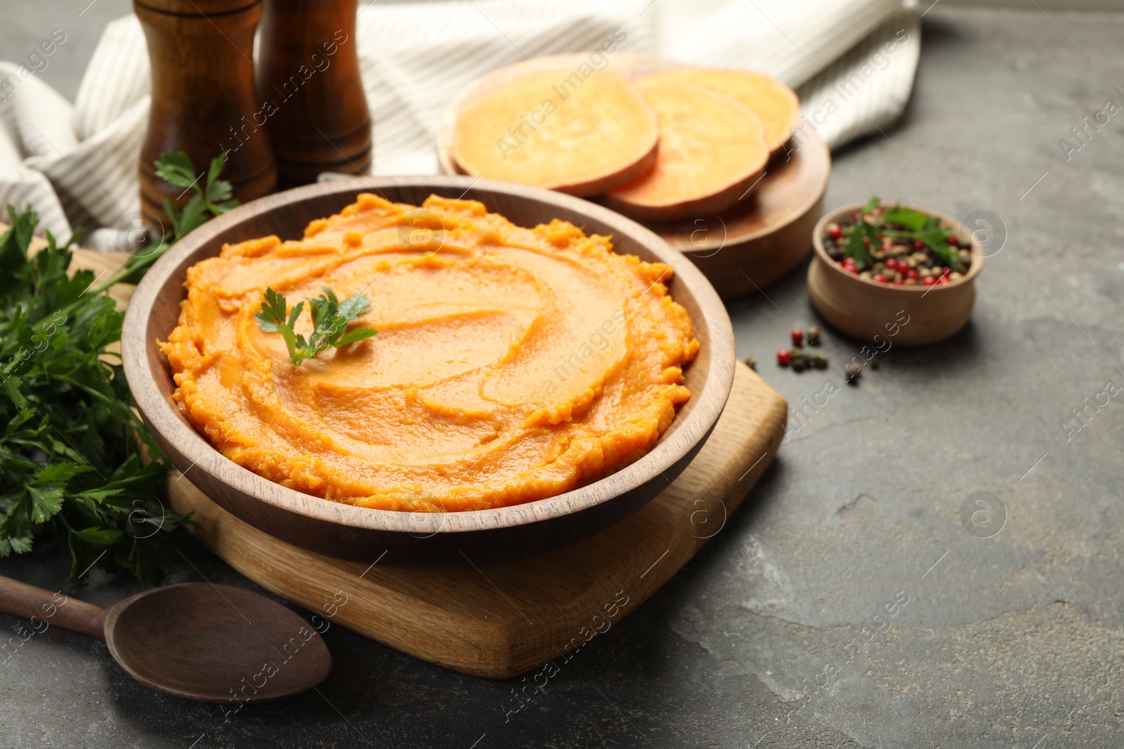 Photo of Delicious mashed sweet potatoes in bowl, pieces of fresh vegetable, spices and spoon on gray textured table, closeup