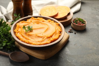 Photo of Delicious mashed sweet potatoes in bowl, pieces of fresh vegetable, spices and spoon on gray textured table, closeup