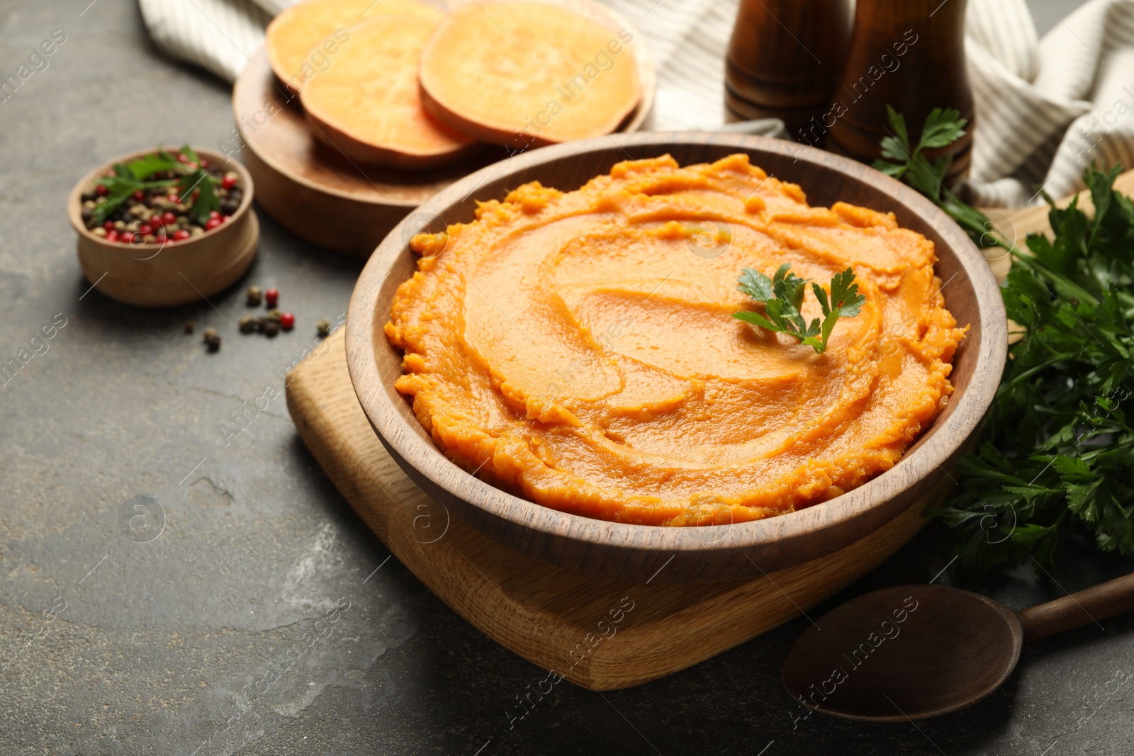 Photo of Delicious mashed sweet potatoes in bowl, pieces of fresh vegetable, spices and spoon on gray textured table, closeup