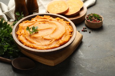 Photo of Delicious mashed sweet potatoes in bowl, pieces of fresh vegetable, spices and spoon on gray textured table, closeup