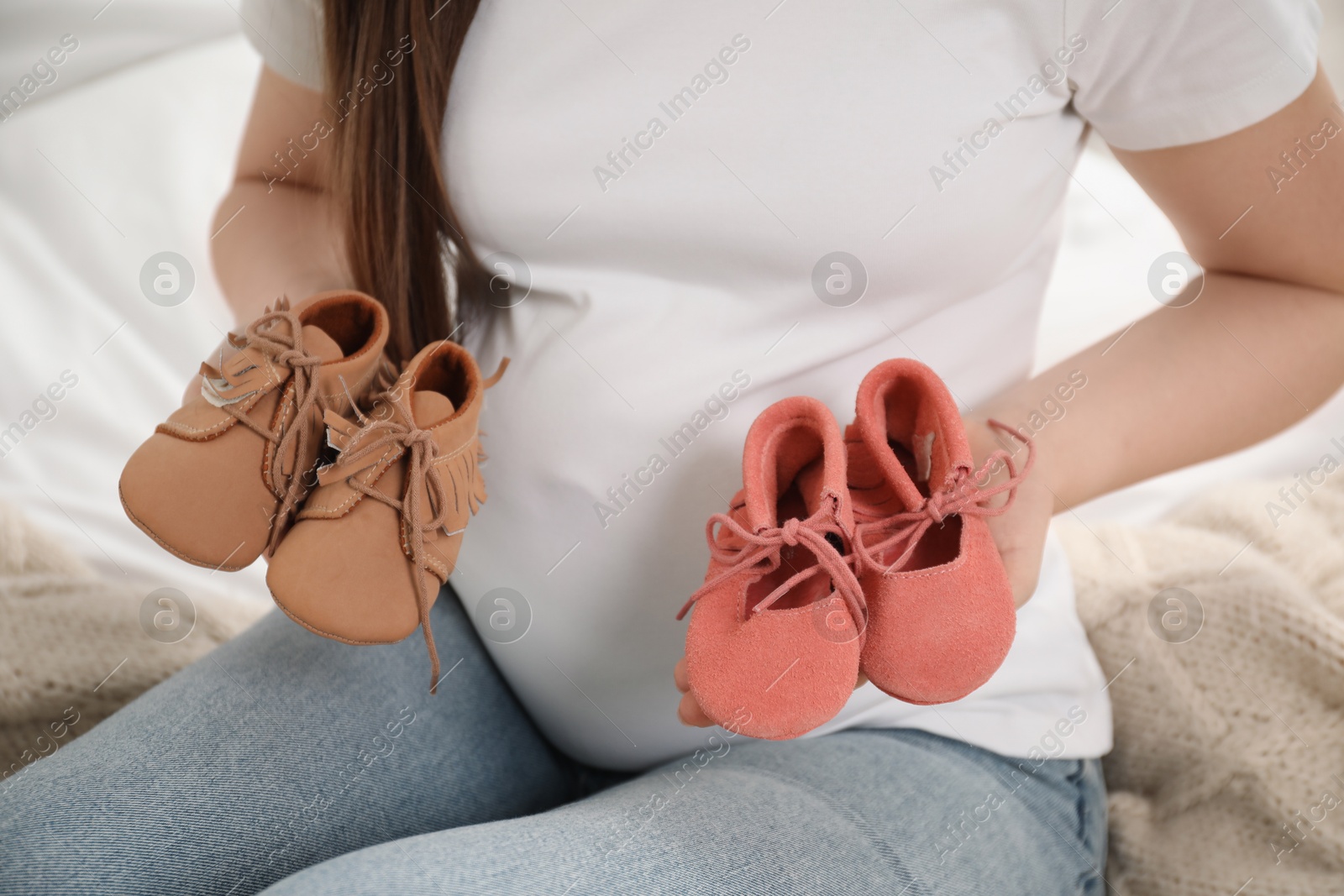 Photo of Expecting twins. Pregnant woman holding two pairs of shoes at home, closeup