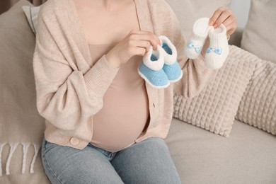 Photo of Expecting twins. Pregnant woman holding two pairs of shoes at home, closeup