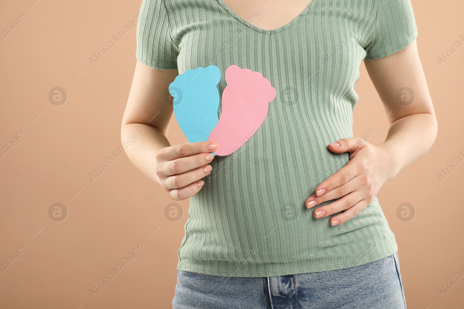 Photo of Expecting twins. Pregnant woman holding two paper cutouts of feet on light brown background, closeup