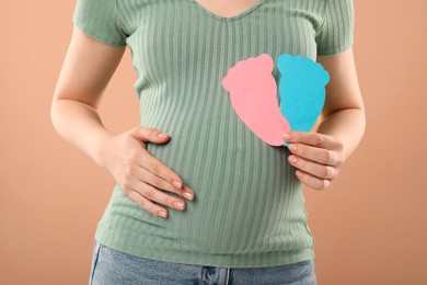 Photo of Expecting twins. Pregnant woman holding two paper cutouts of feet on light brown background, closeup