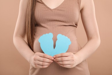 Photo of Expecting twins. Pregnant woman holding two paper cutouts of feet on light brown background, closeup