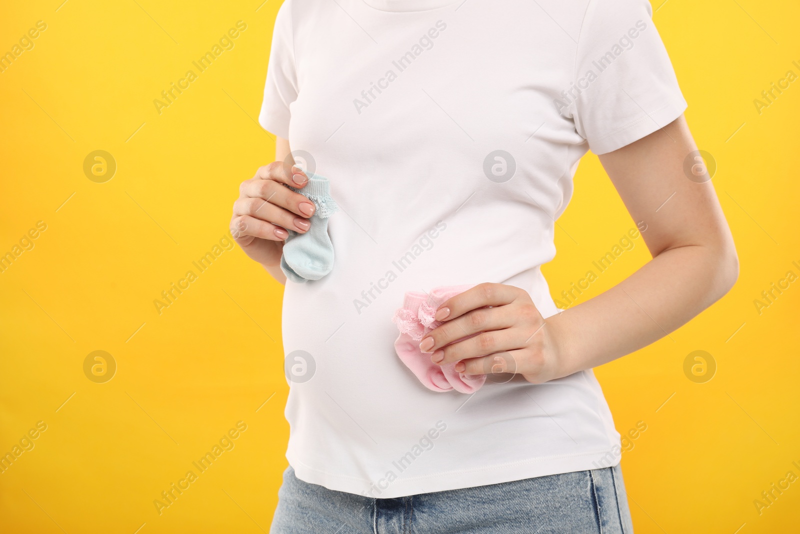 Photo of Expecting twins. Pregnant woman holding two pairs of socks on yellow background, closeup