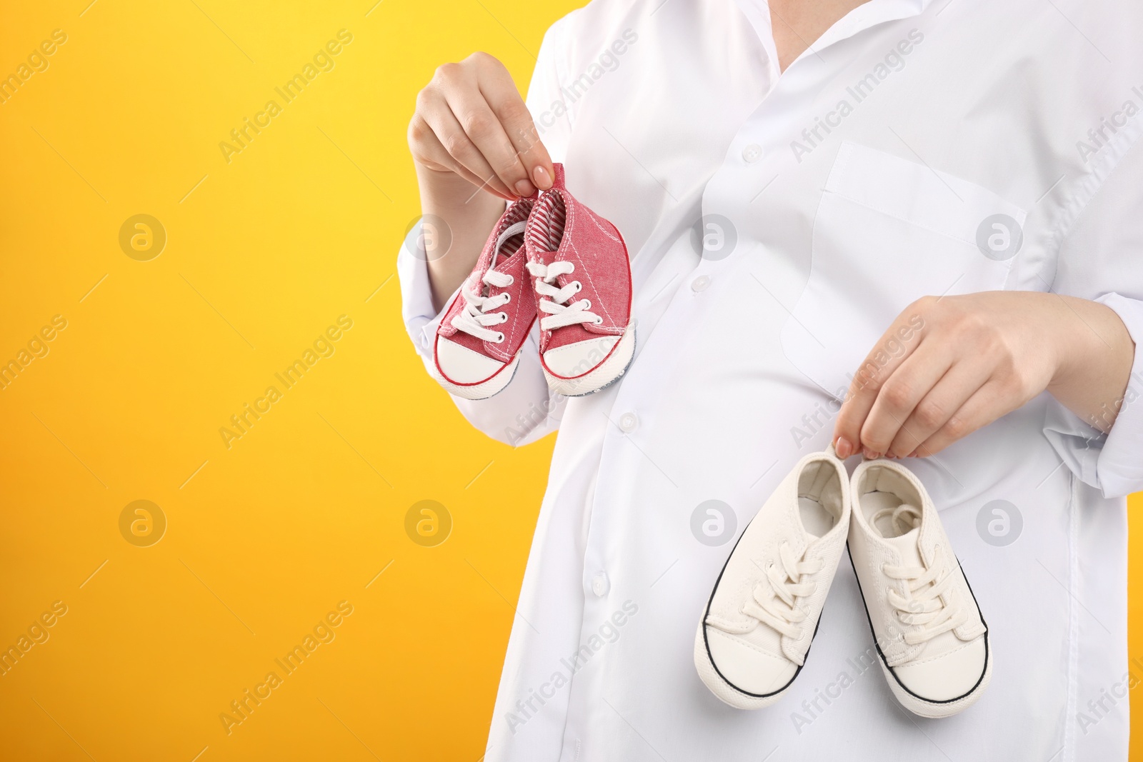 Photo of Expecting twins. Pregnant woman holding two pairs of shoes on yellow background, closeup. Space for text