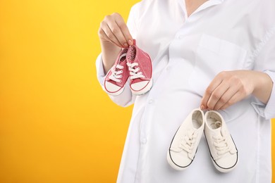 Expecting twins. Pregnant woman holding two pairs of shoes on yellow background, closeup. Space for text