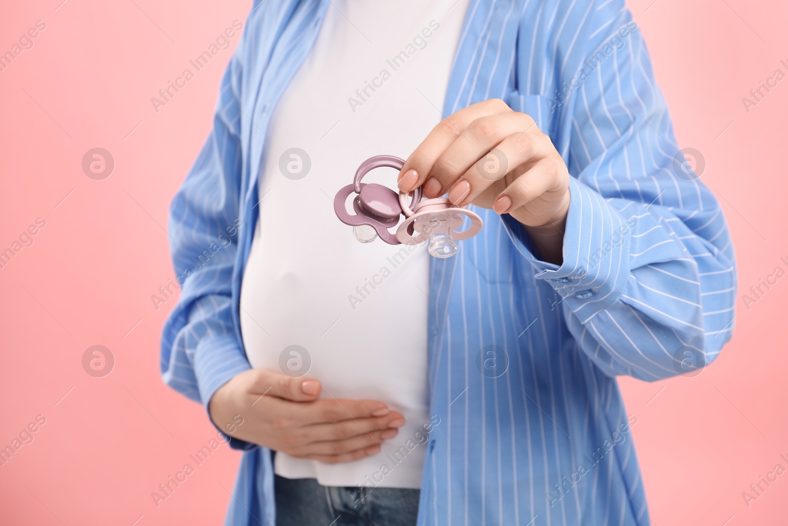 Photo of Expecting twins. Pregnant woman holding two pacifiers on pink background, closeup