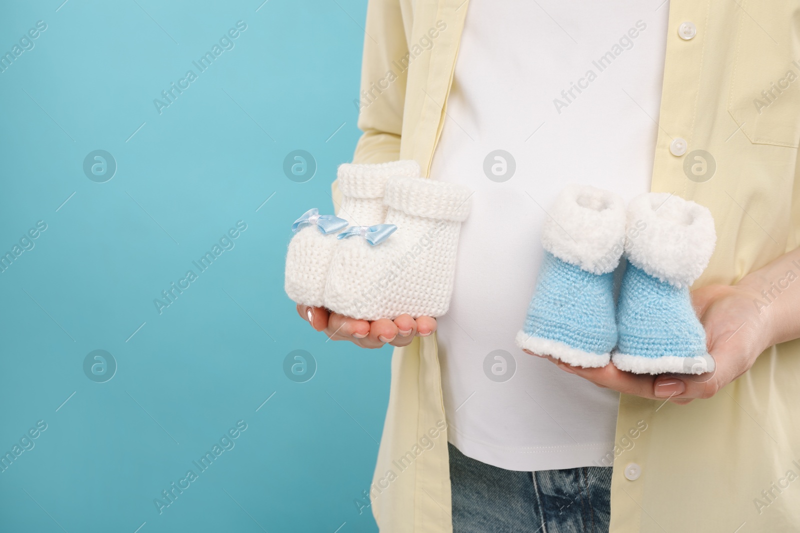 Photo of Expecting twins. Pregnant woman holding two pairs of baby shoes on light blue background, closeup. Space for text