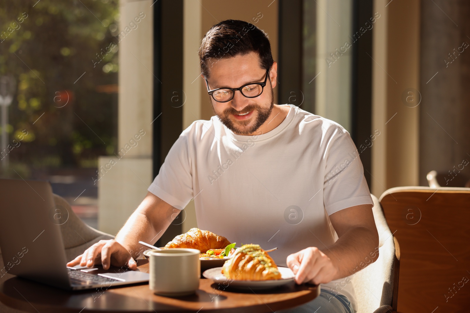 Photo of Happy man having tasty breakfast and using laptop in cafe