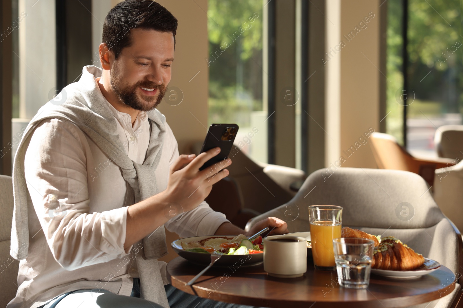 Photo of Happy man having tasty breakfast and using smartphone in cafe