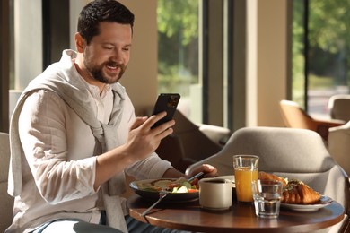 Happy man having tasty breakfast and using smartphone in cafe