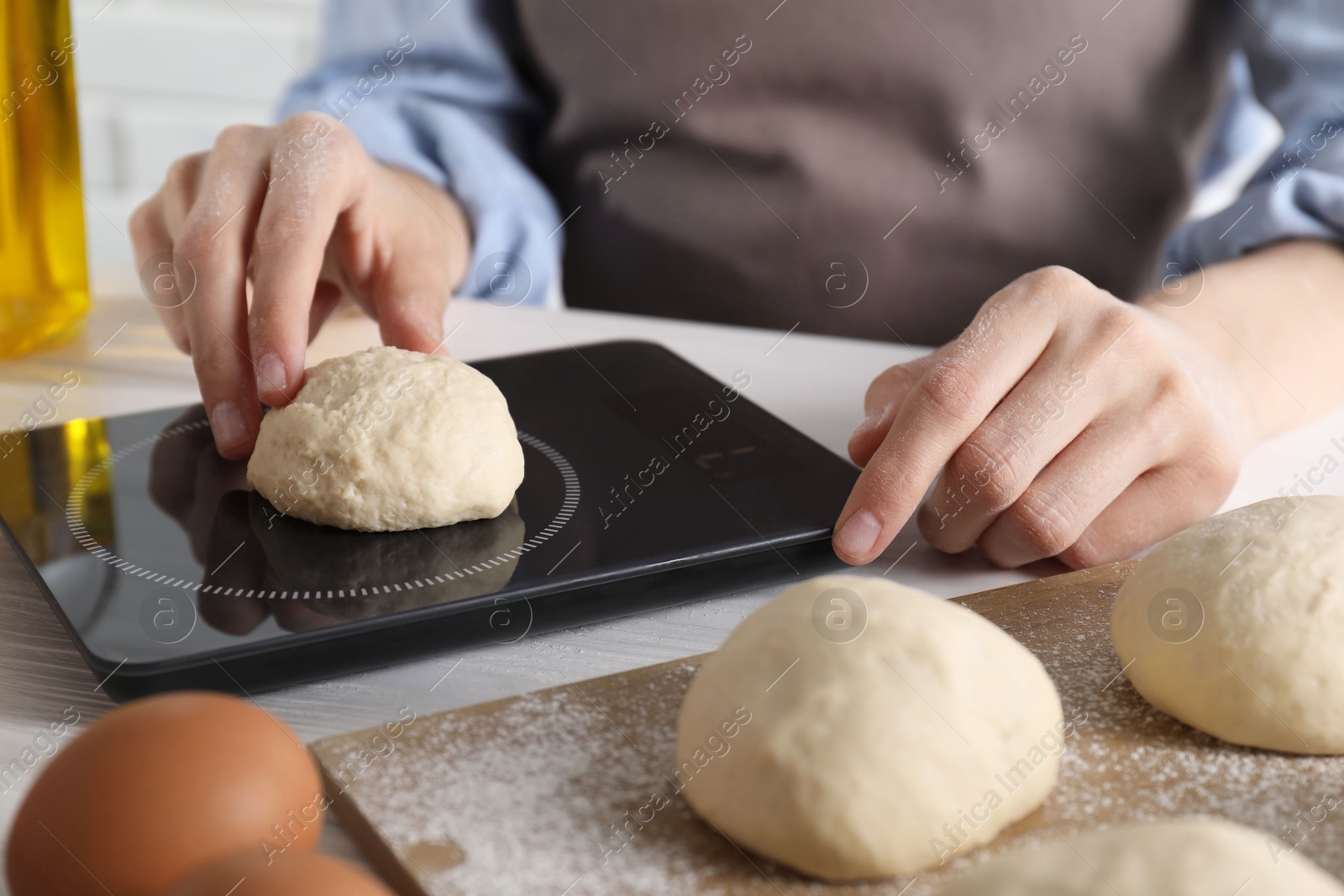 Photo of Woman weighing raw dough ball on kitchen scale at white wooden table, closeup