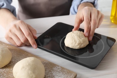 Photo of Woman weighing raw dough ball on kitchen scale at white wooden table, closeup