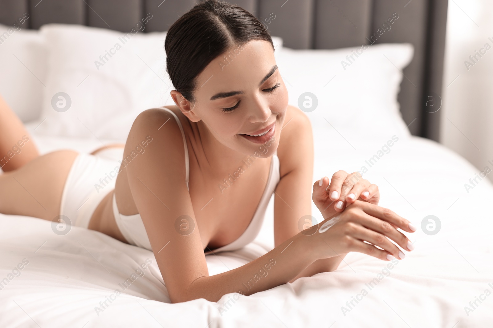 Photo of Smiling woman applying cream onto hand on bed. Body care