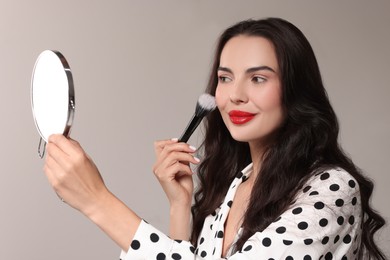 Beautiful woman with mirror applying makeup on light grey background