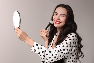 Smiling woman with mirror applying makeup on light grey background