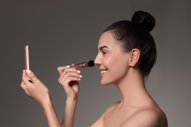 Smiling woman with mirror applying makeup on grey background