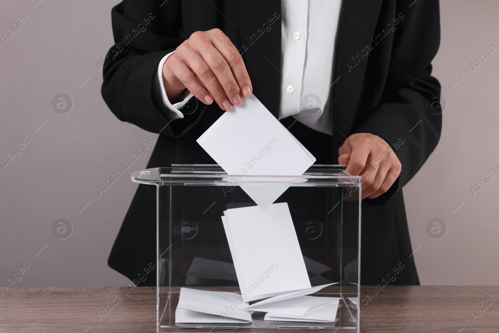 Photo of Woman putting her vote into ballot box at wooden table against grey background, closeup