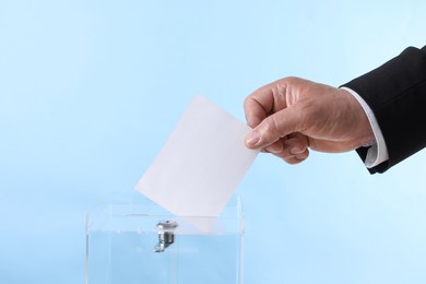 Man putting his vote into ballot box against light blue background, closeup