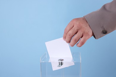 Man putting his vote into ballot box against light blue background, closeup