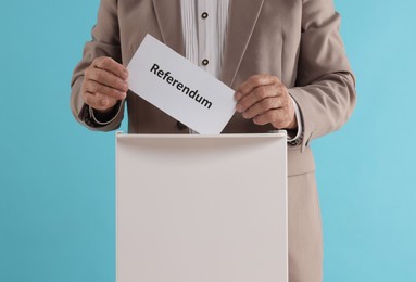 Referendum. Man putting his vote into ballot box at wooden table against light blue background, closeup