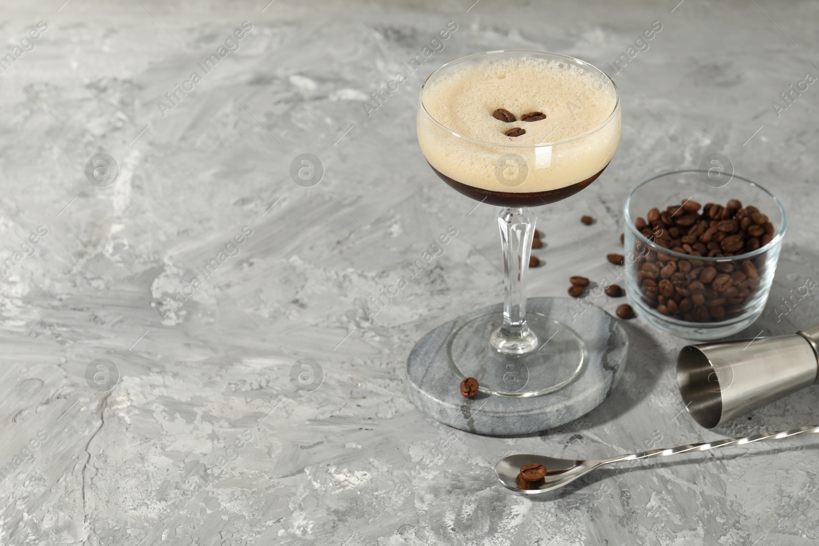 Photo of Glass of fresh coffee cocktail, beans in bowl and bartender equipment on gray textured table, space for text