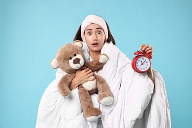 Photo of Overslept young woman with teddy bear and alarm clock wrapped in blanket on light blue background