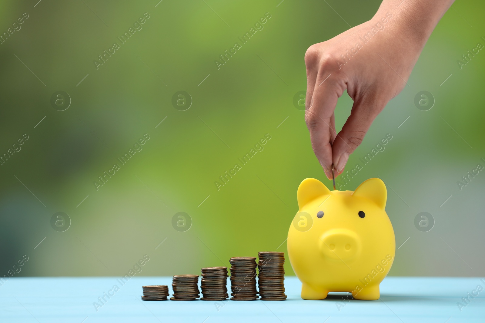 Photo of Woman putting coin into yellow piggy bank at light blue table against blurred background, closeup. Space for text