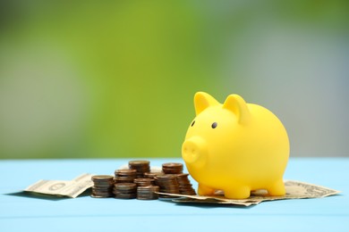 Photo of Yellow piggy bank, dollar banknotes and stacks of coins on light blue table against blurred background