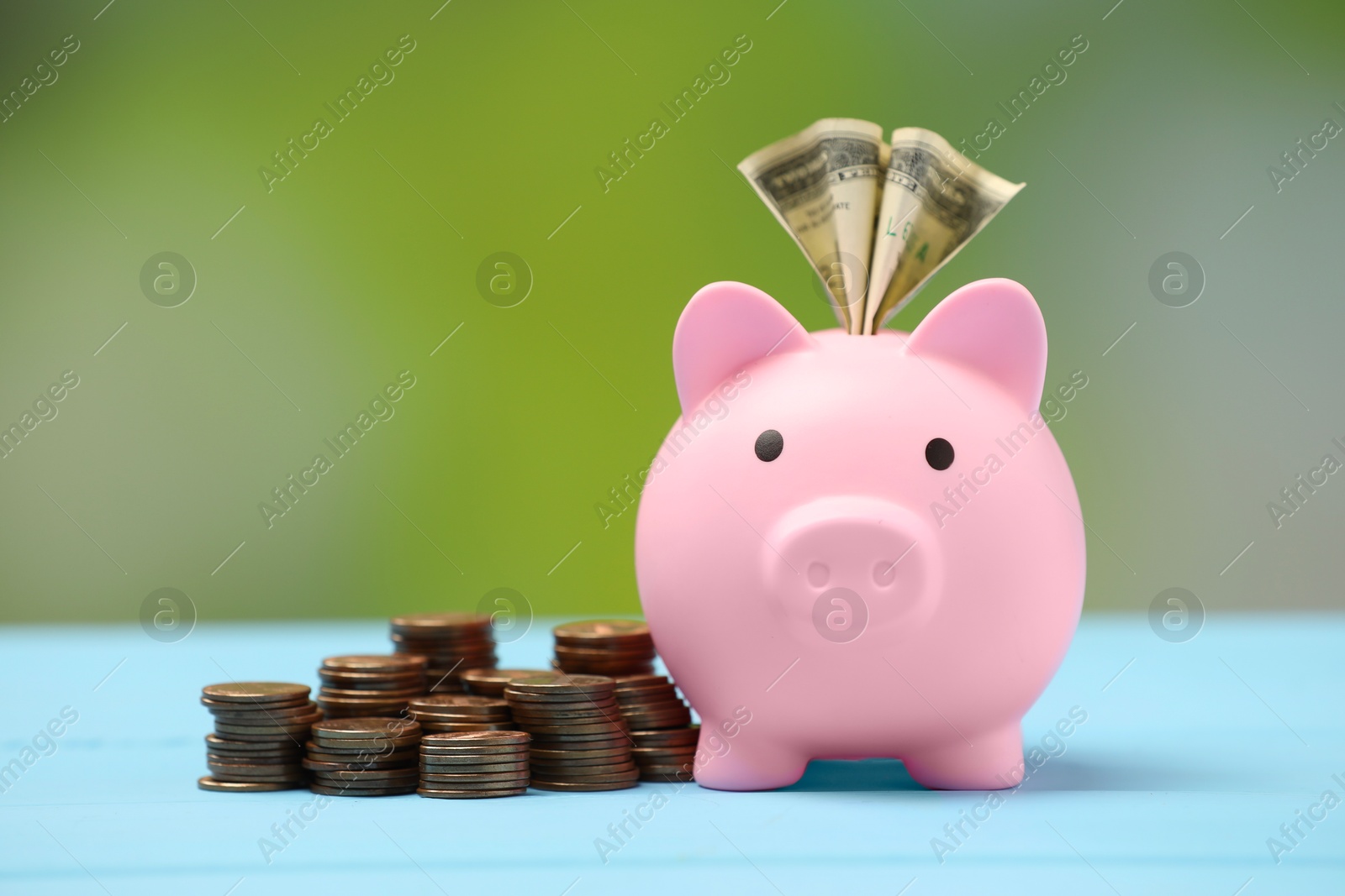 Photo of Pink piggy bank with dollar banknote and stacks of coins on light blue table against blurred background, closeup