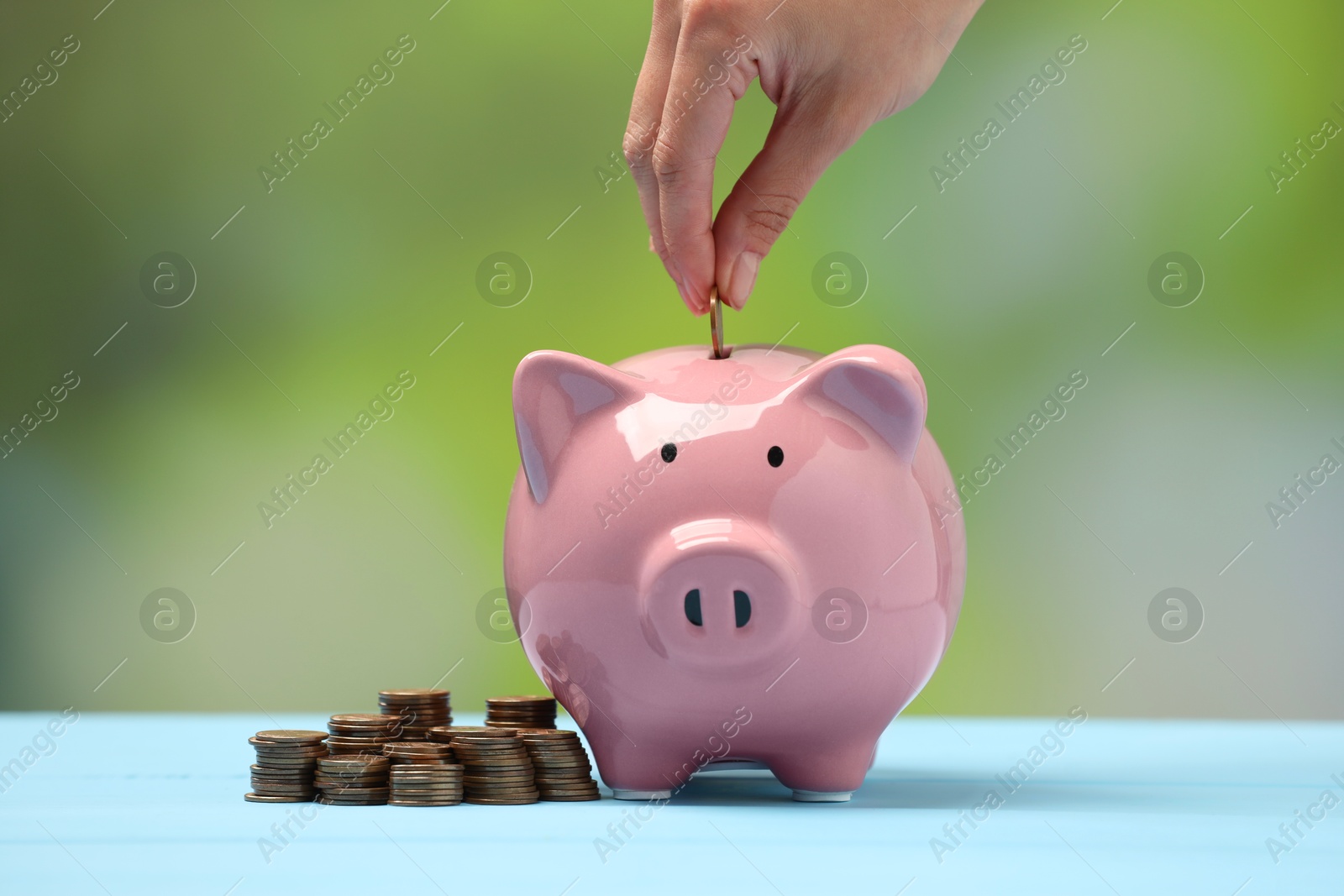 Photo of Woman putting coin into pink piggy bank at light blue table against blurred background, closeup