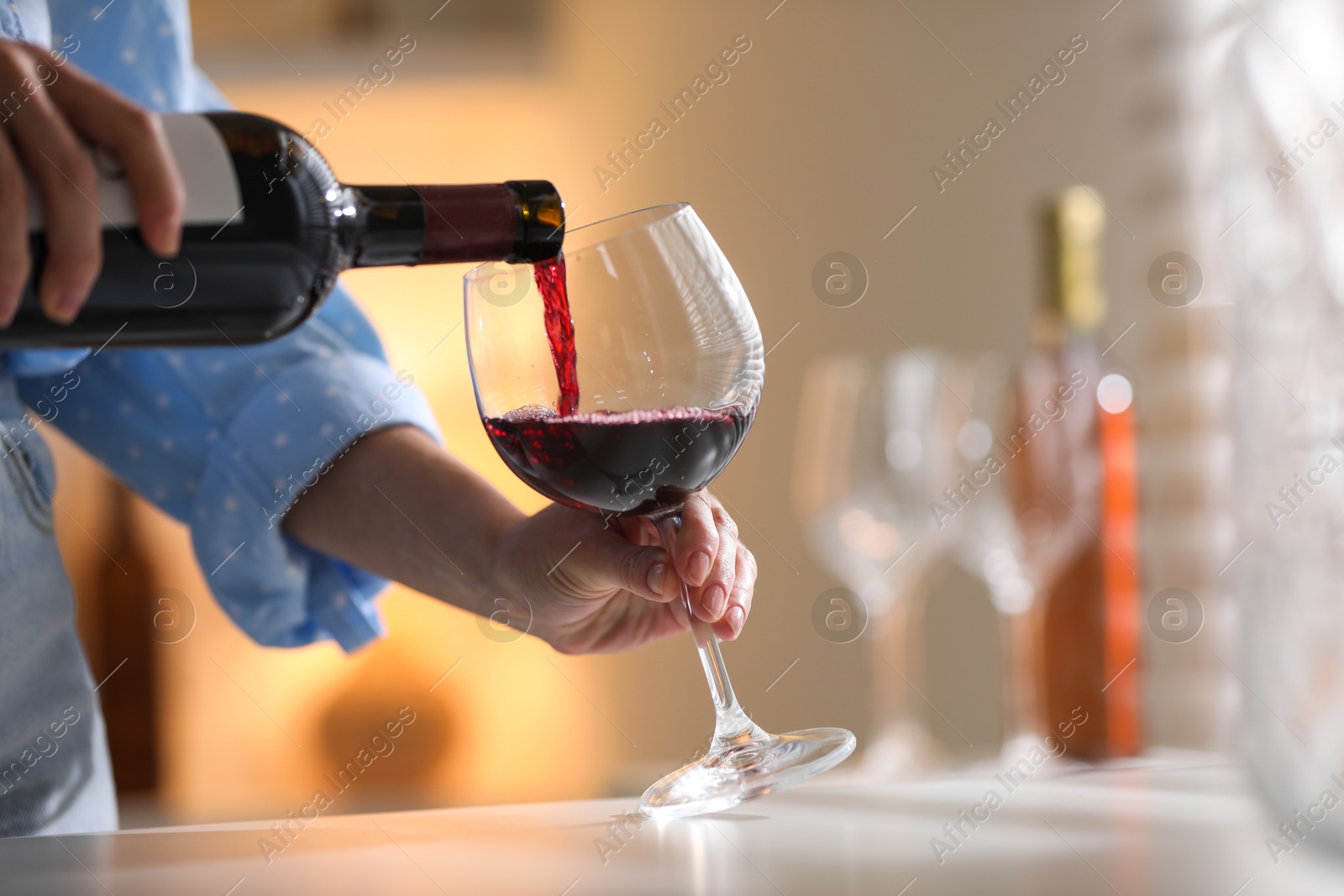 Photo of Woman pouring red wine into glass at white table indoors, closeup